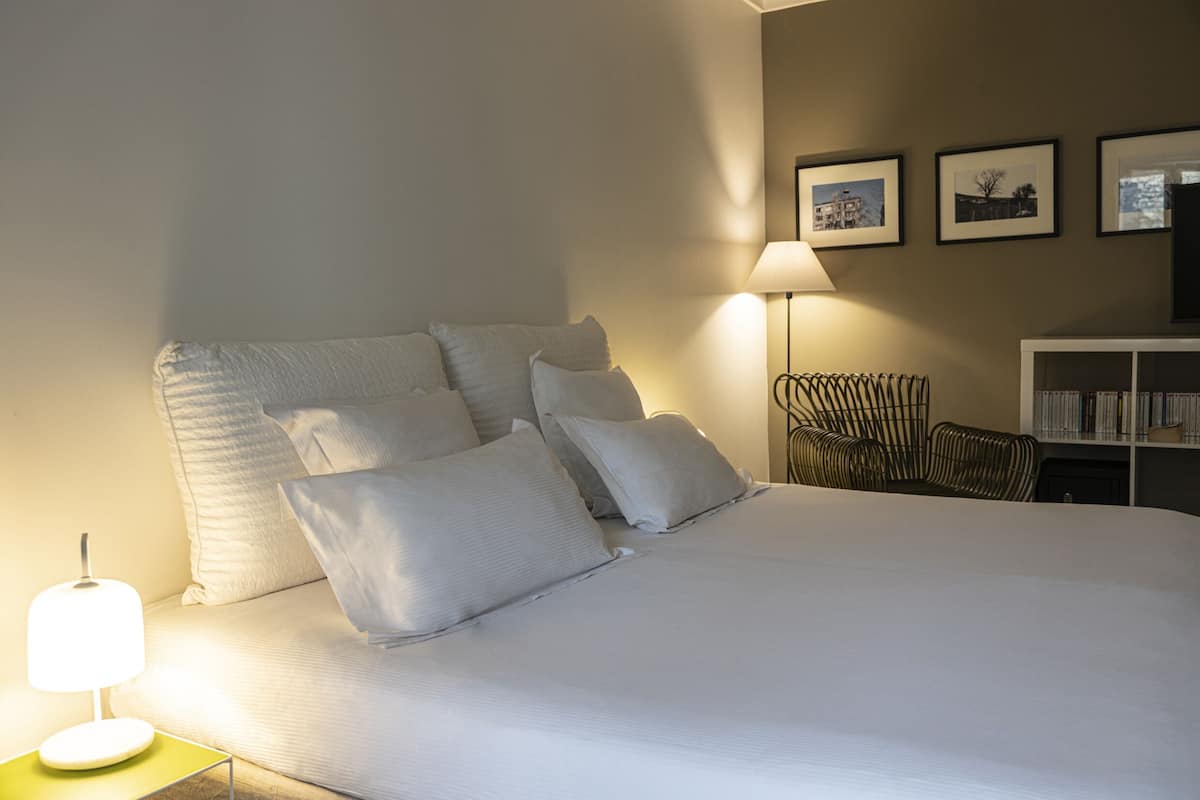Una cama bien hecha con sábanas y almohadas blancas, una lámpara de noche, una silla y una estantería con libros.