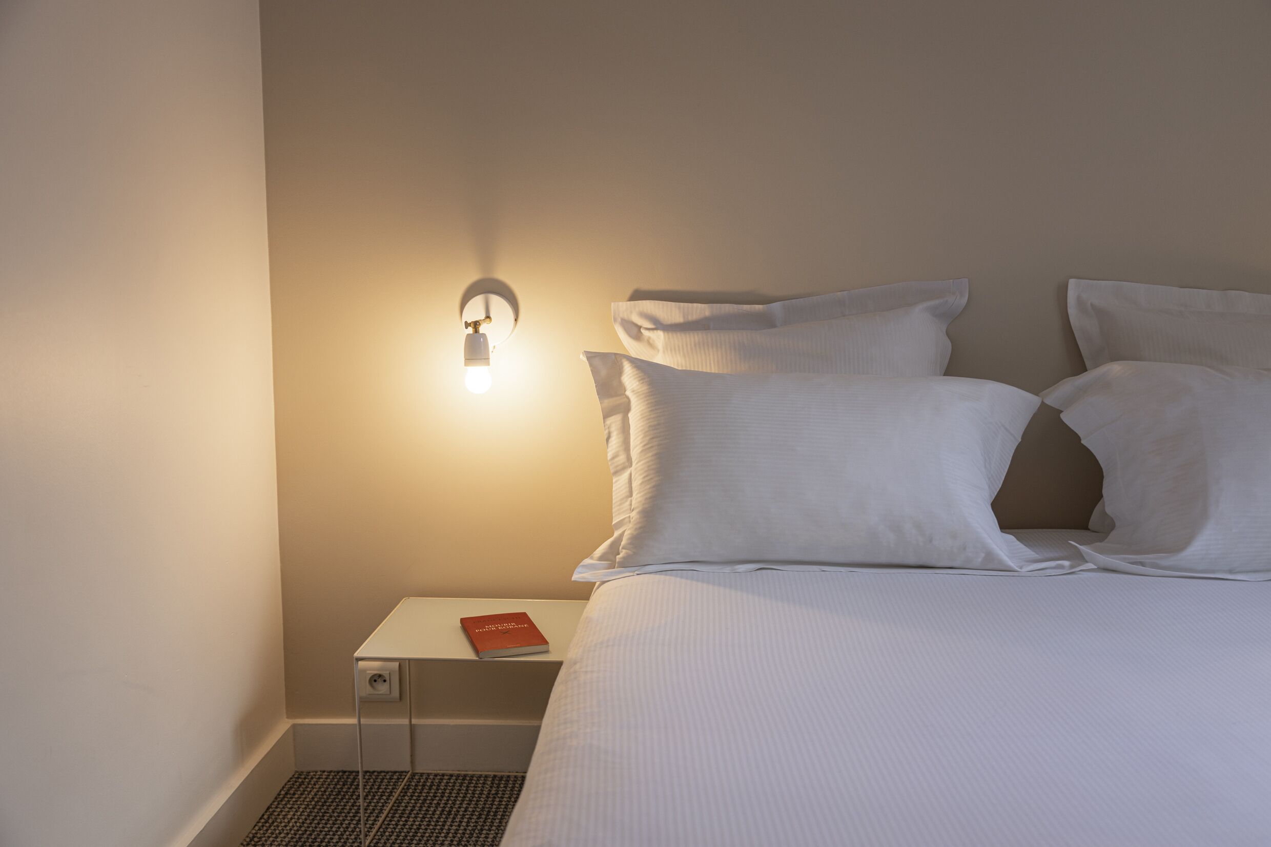 A neatly made bed with white linens and pillows, a bedside table with a book, and a wall-mounted light fixture.