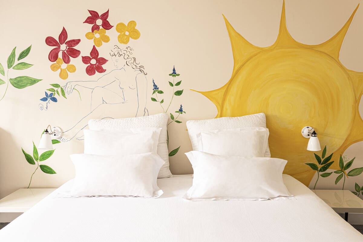 Una cama con ropa blanca y almohadas, una pared con un mural de sol y adornos florales, y una mesita de noche con una lámpara.