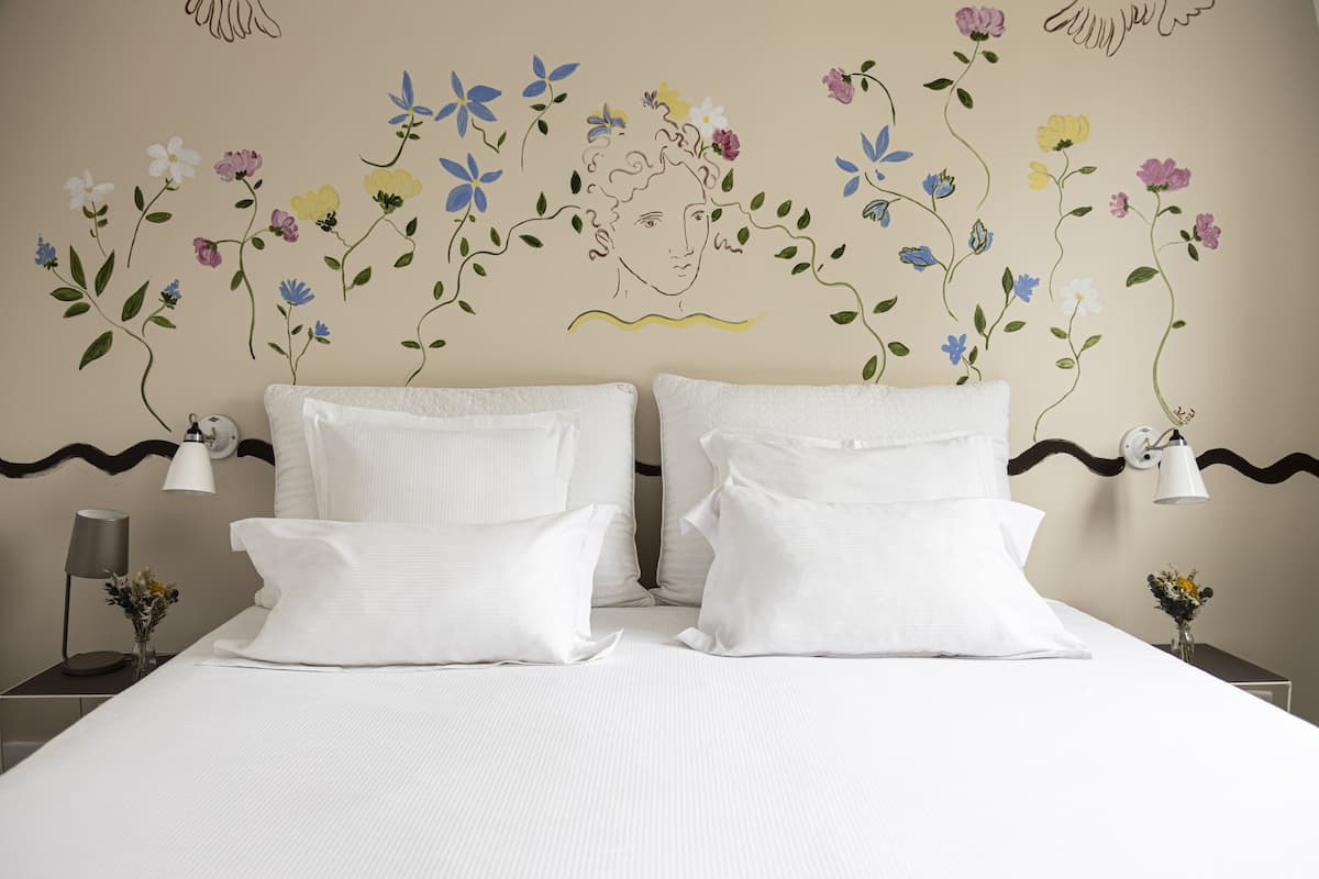 Habitación de hotel con cama, almohadas blancas, un mural floral y una mesita de noche con lámpara y un florero.