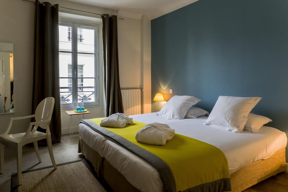 Una habitación de hotel con una cama, una silla, una ventana con cortinas, una mesita de noche y una lámpara.