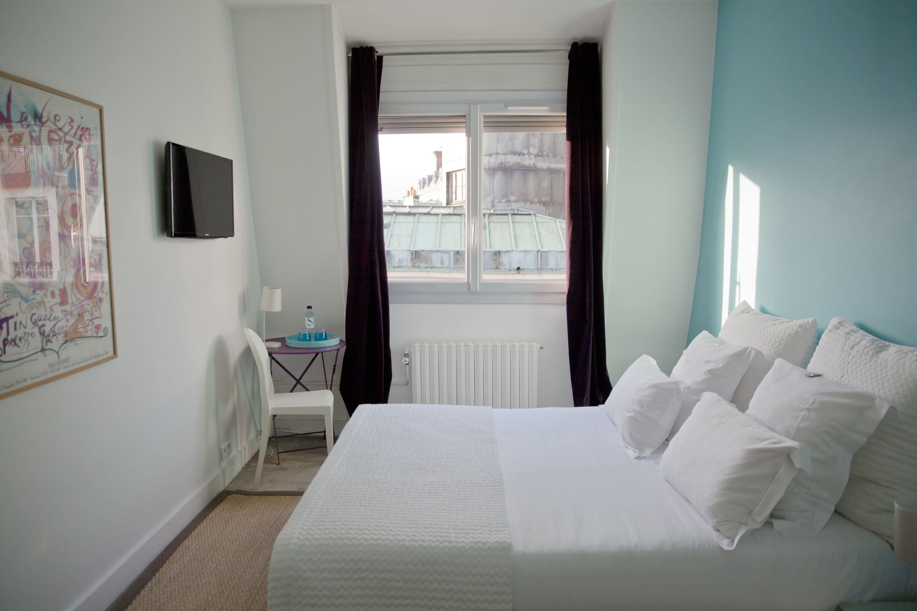 A hotel room with a bed, a desk with a chair, a window with curtains, a radiator, and a wall-mounted TV.
