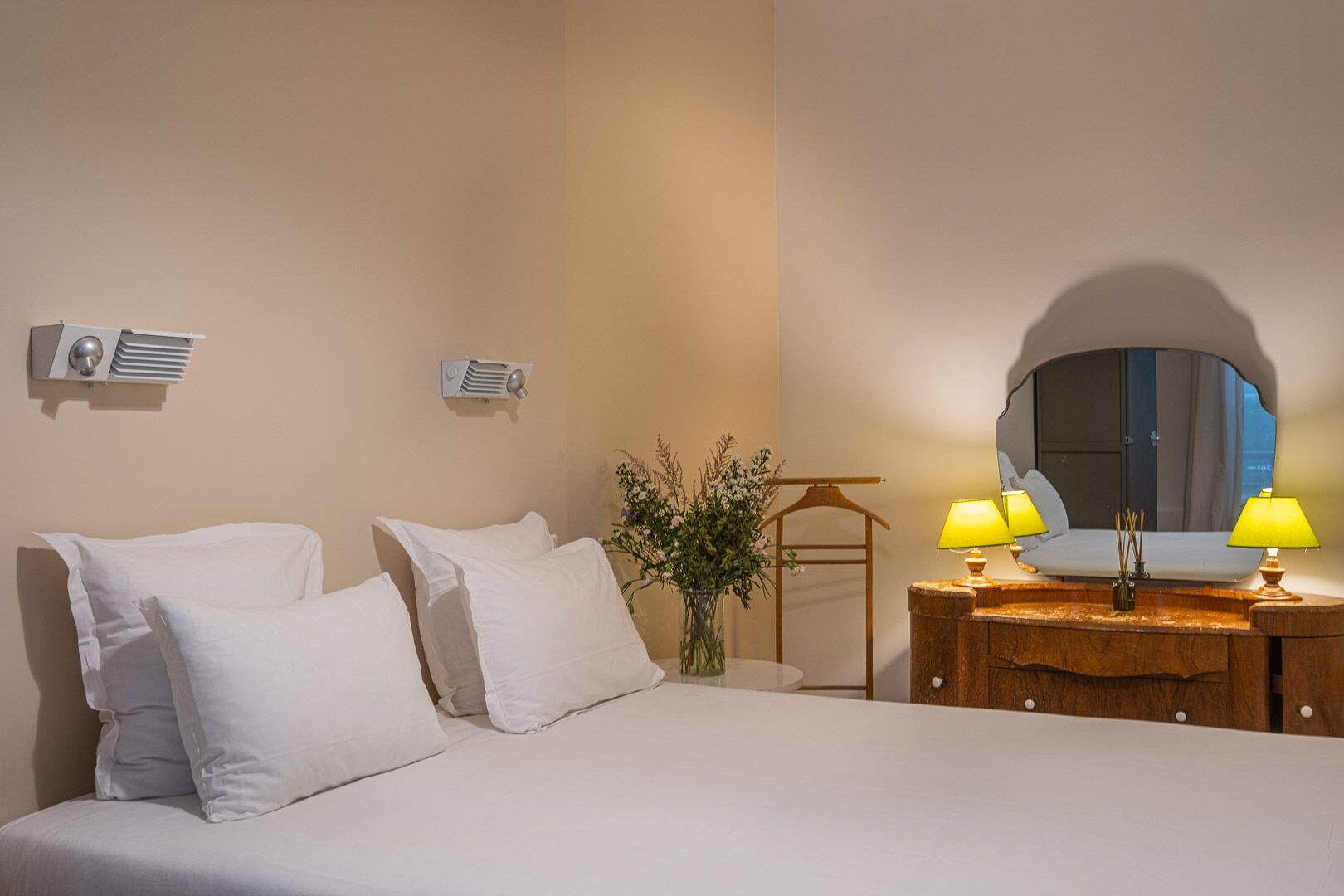 Hotellihuone, jossa on sänky, yöpöytälamput, peili, lipasto ja pieni kasvi.