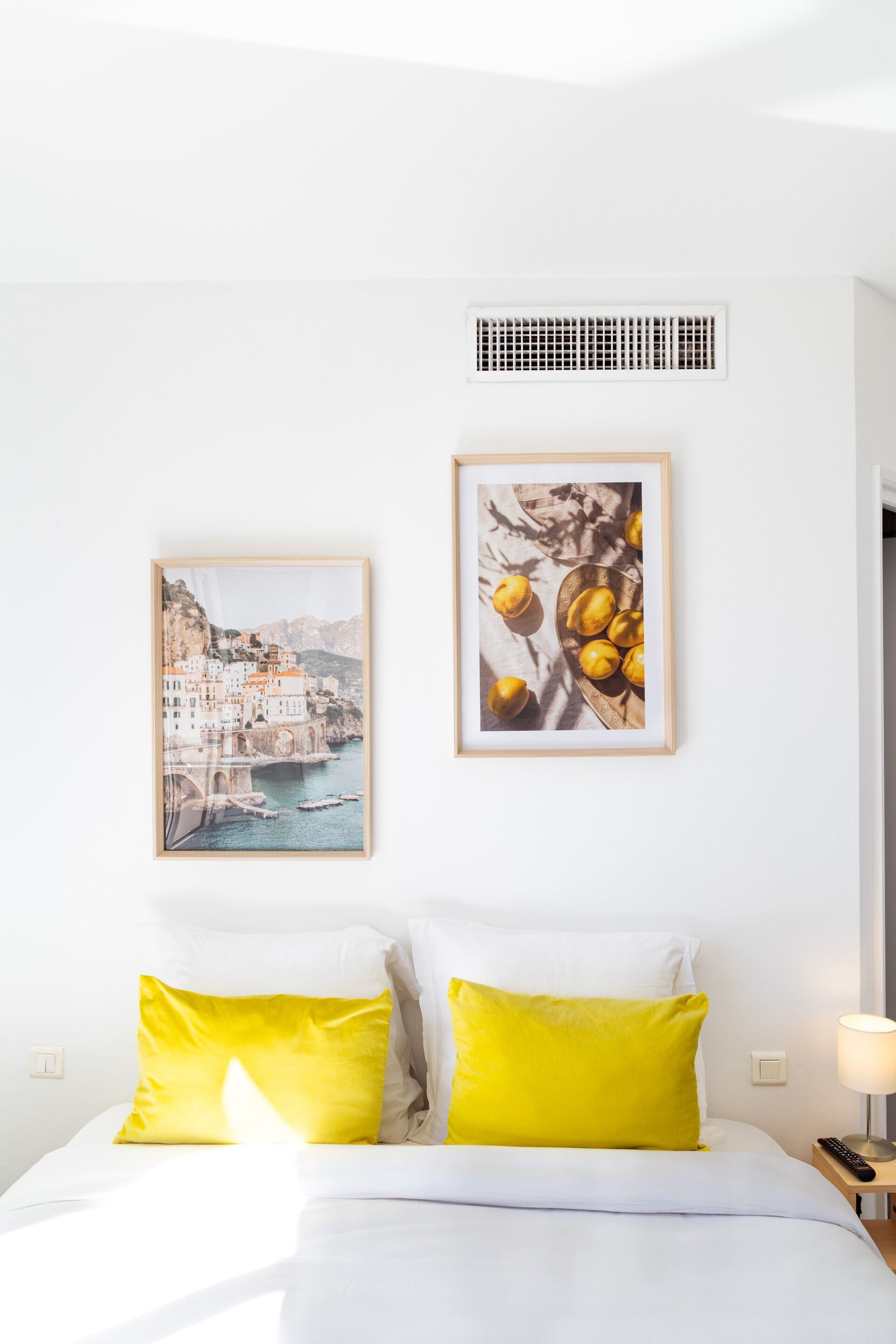 Een hotelkamer met een bed, twee kussens, een lijst met een foto van een kustplaats en een lijst met citroenen.