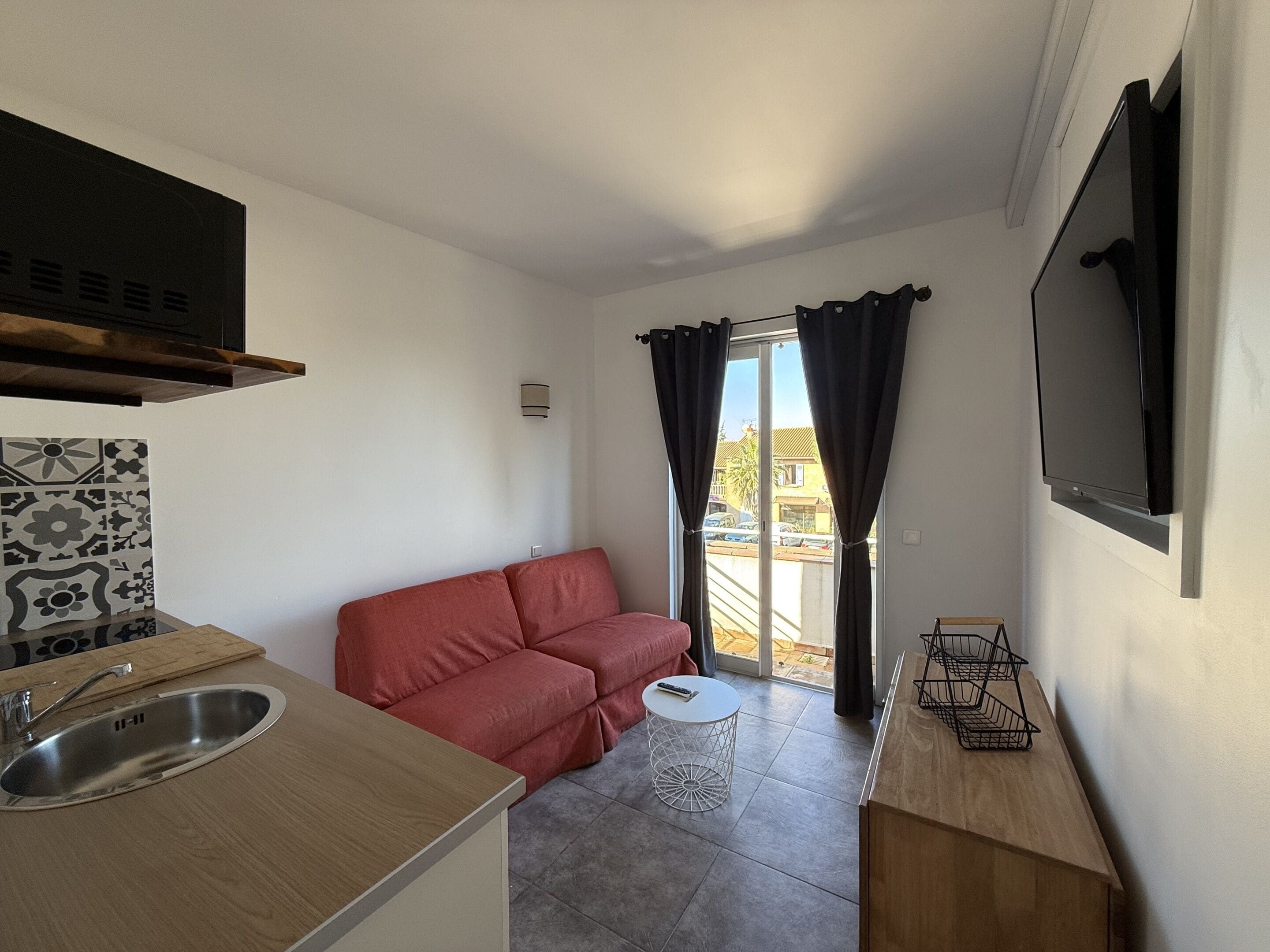 A compact kitchen with a sink, a red sofa, a small round table, and a TV mounted on the wall.