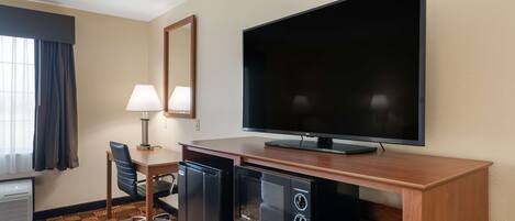 A hotel room with a flat-screen TV, a mini-fridge, a microwave, a desk with a chair, and a window with curtains.