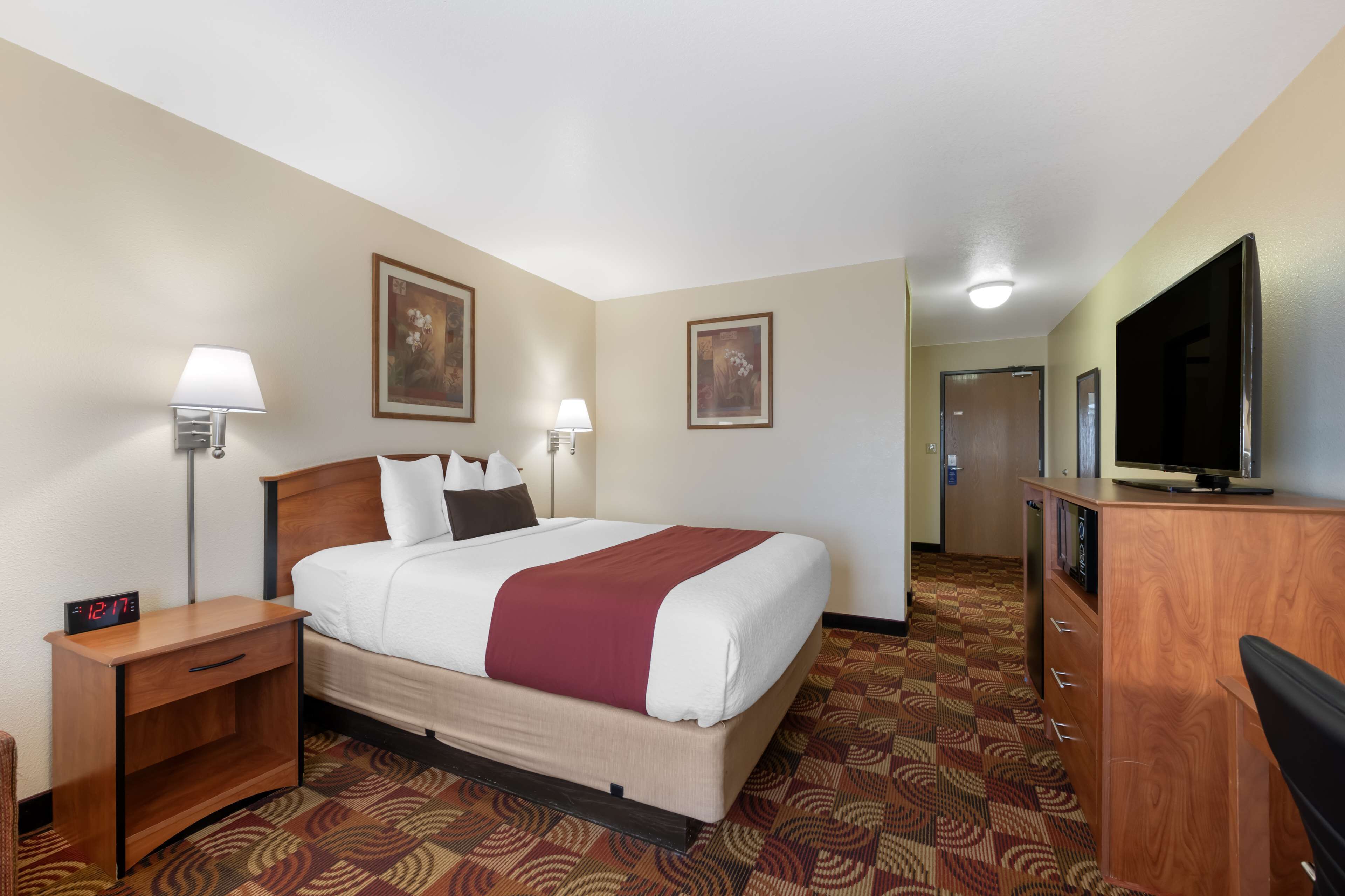 A hotel room with a bed, bedside lamps, a desk, a television, and a carpeted floor.