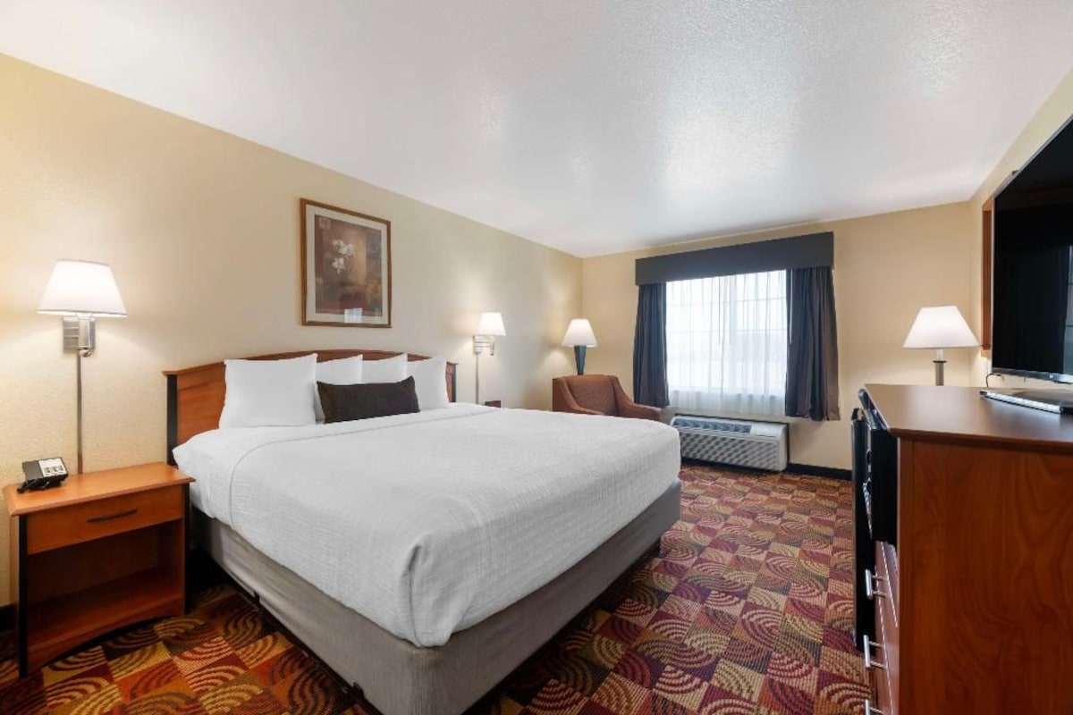 A hotel room with a large bed, two bedside tables with lamps, a desk with a chair, a television, and a window with curtains.