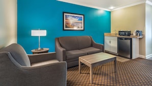 A modern hotel room with a teal accent wall, a gray sofa, a small table, a lamp, and a kitchenette area. - Best Western Ocean City Hotel & Suites (Ocean City)