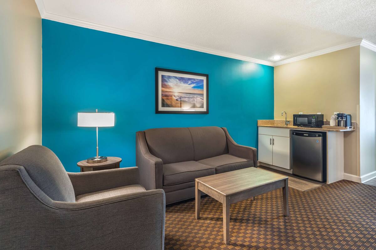 A modern hotel room with a teal accent wall, a gray sofa, a small table, a lamp, and a kitchenette area.