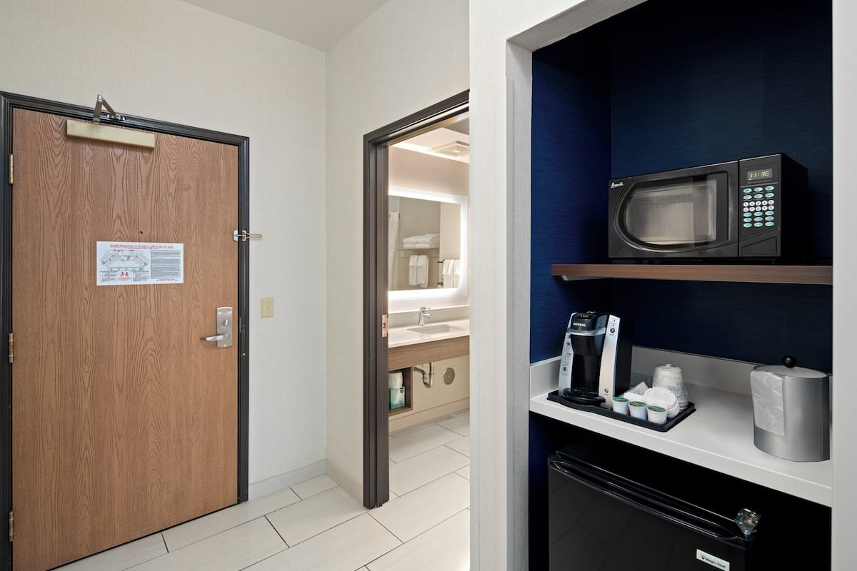 A hotel room with a wooden door, a visible emergency evacuation plan, a microwave on a shelf, a coffee maker, and a trash bin.