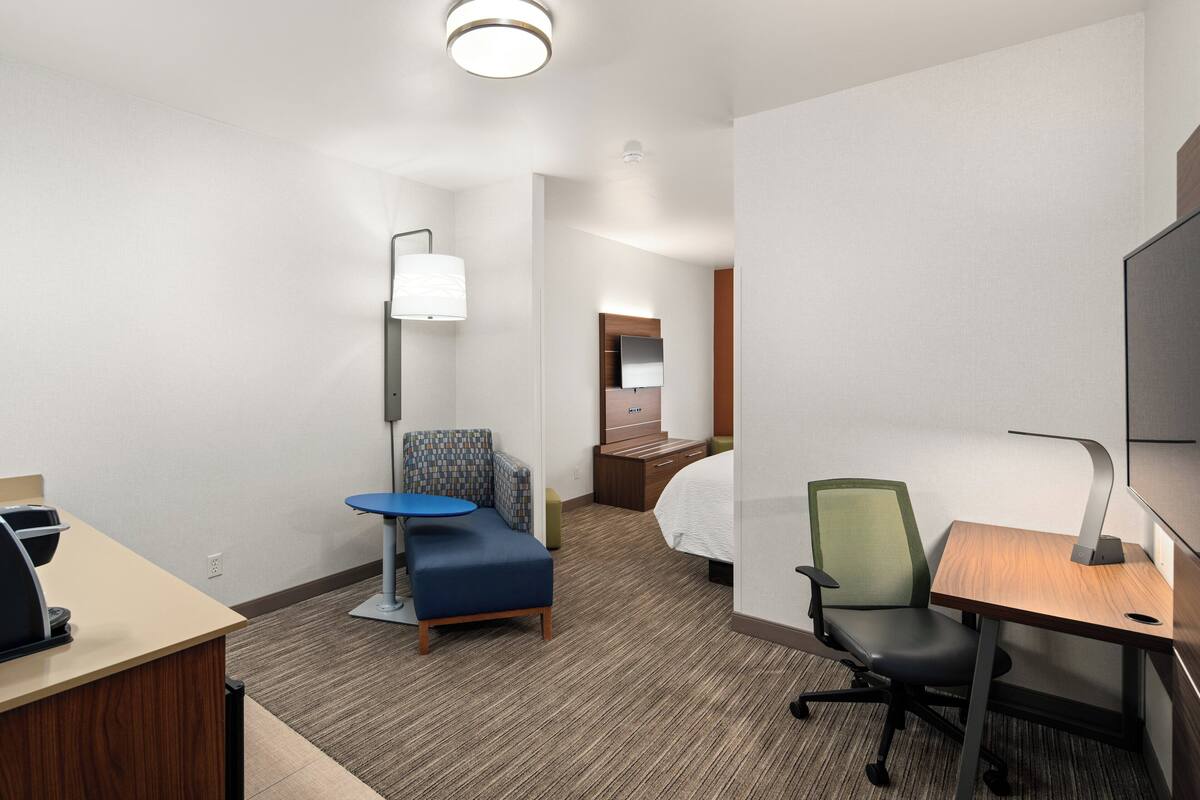 A hotel room with a desk, a blue chair, a green chair, a bed, and a TV.