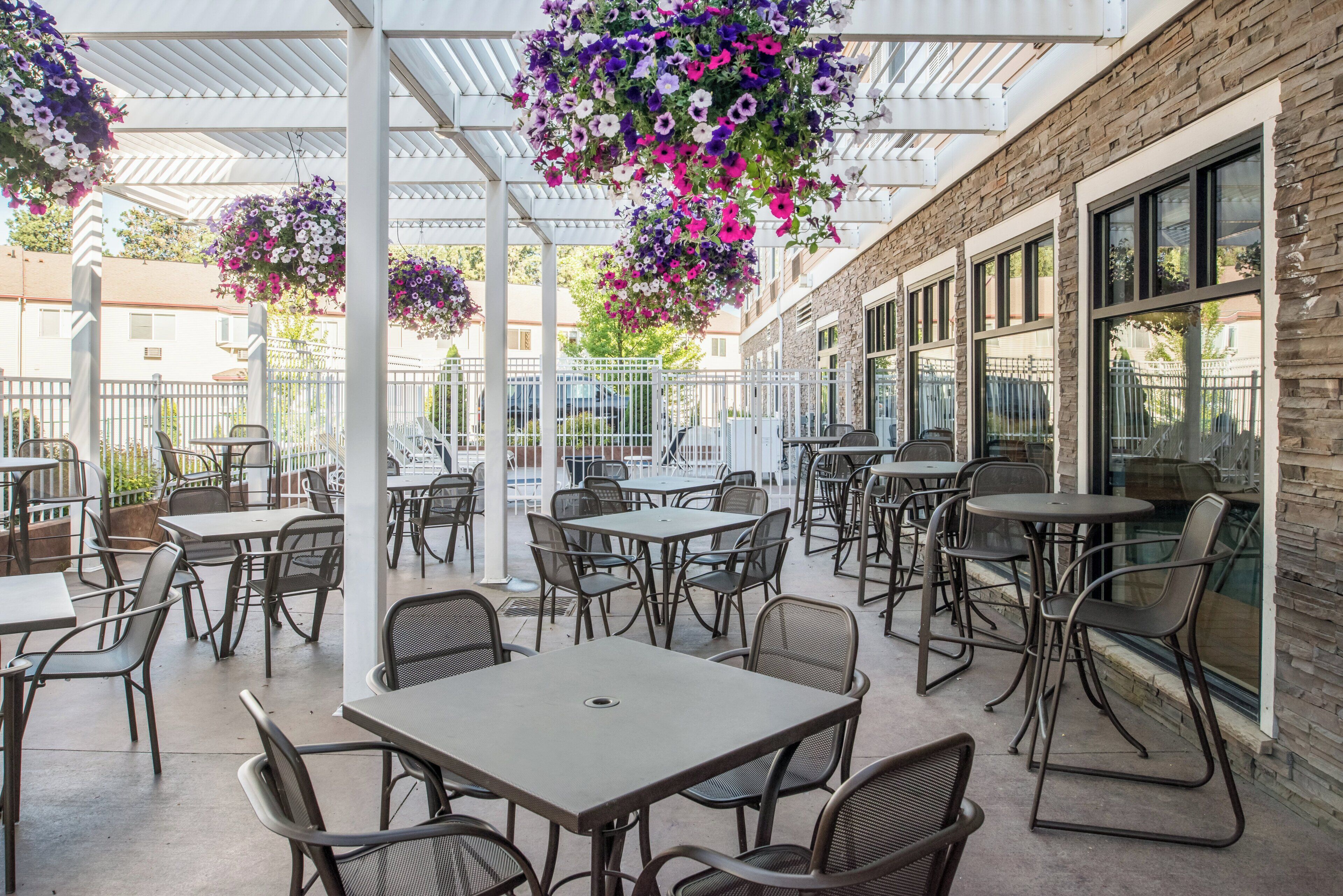 Una zona patio all'aperto con tavoli e sedie, una sezione coperta con cesti di fiori pensili e un edificio con ampie finestre.