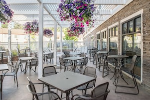 Un patio al aire libre con mesas y sillas, una zona cubierta con cestas de flores colgantes y un edificio con amplias ventanas.
