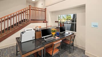 Un centro de negocios con una escalera de madera, dos escritorios con computadoras, sillas y un ventanal con vistas al estacionamiento.