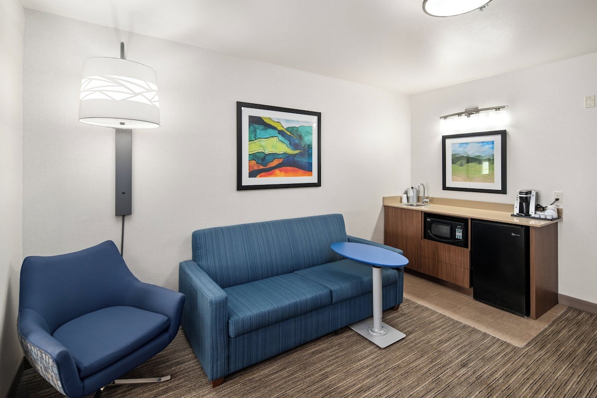 A modern hotel room with a blue sofa, a small round table, a microwave, and a dishwasher. There are two framed artworks on the walls.