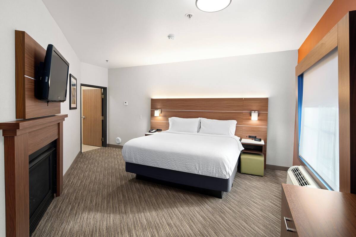 A hotel room with a large bed, a flat-screen TV mounted on the wall, a bedside table with a lamp, and a window with a view.