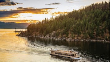 Un bateau à vapeur navigue sur une rivière paisible, bordée d’une forêt, avec un coucher de soleil en arrière-plan.