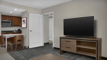 A hotel room with a kitchenette, a flat-screen TV, a seating area with a coffee table, and a carpeted floor.