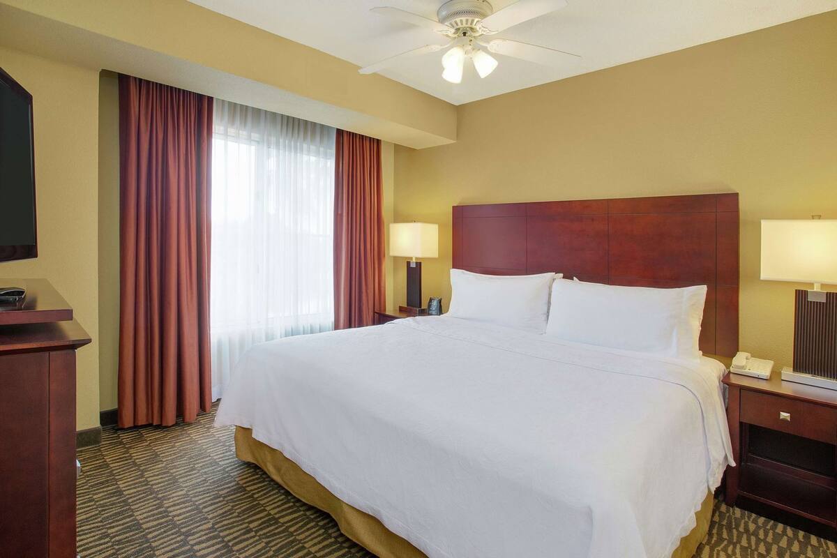 Une chambre d’hôtel avec un grand lit, deux tables de chevet, un bureau avec un téléphone, un ventilateur de plafond et une fenêtre avec des rideaux.