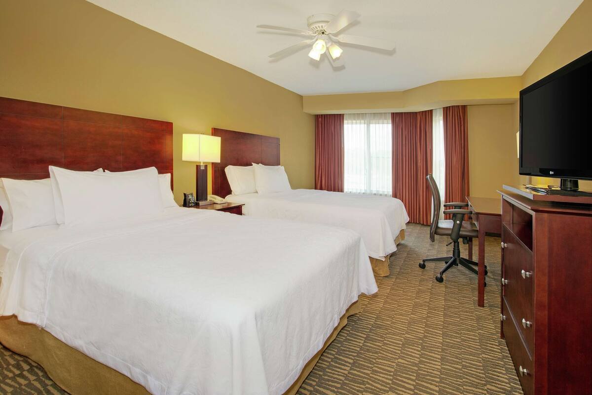 Une chambre d’hôtel avec deux lits, un ventilateur de plafond, une télévision, un bureau avec une chaise et une commode.