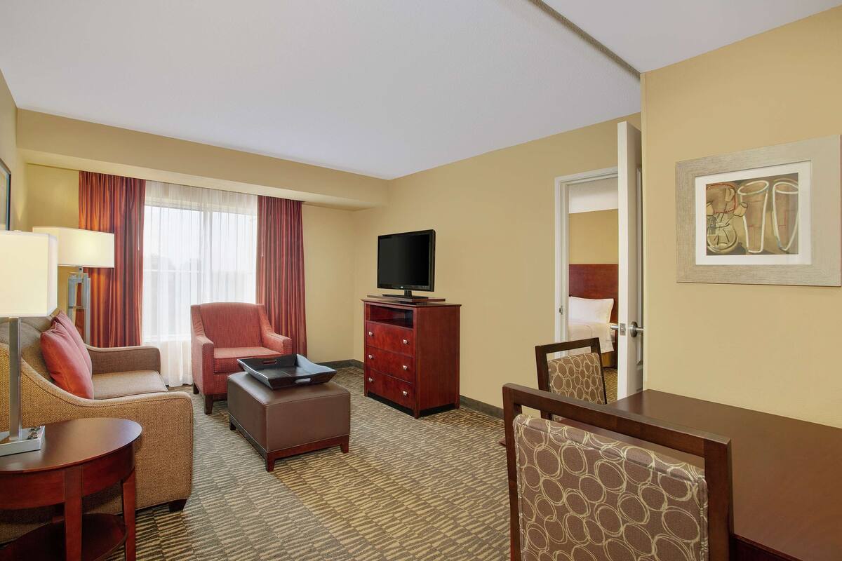 Une chambre d’hôtel comprenant un canapé, un fauteuil, un pouf, un meuble pour la télévision et une table à manger.