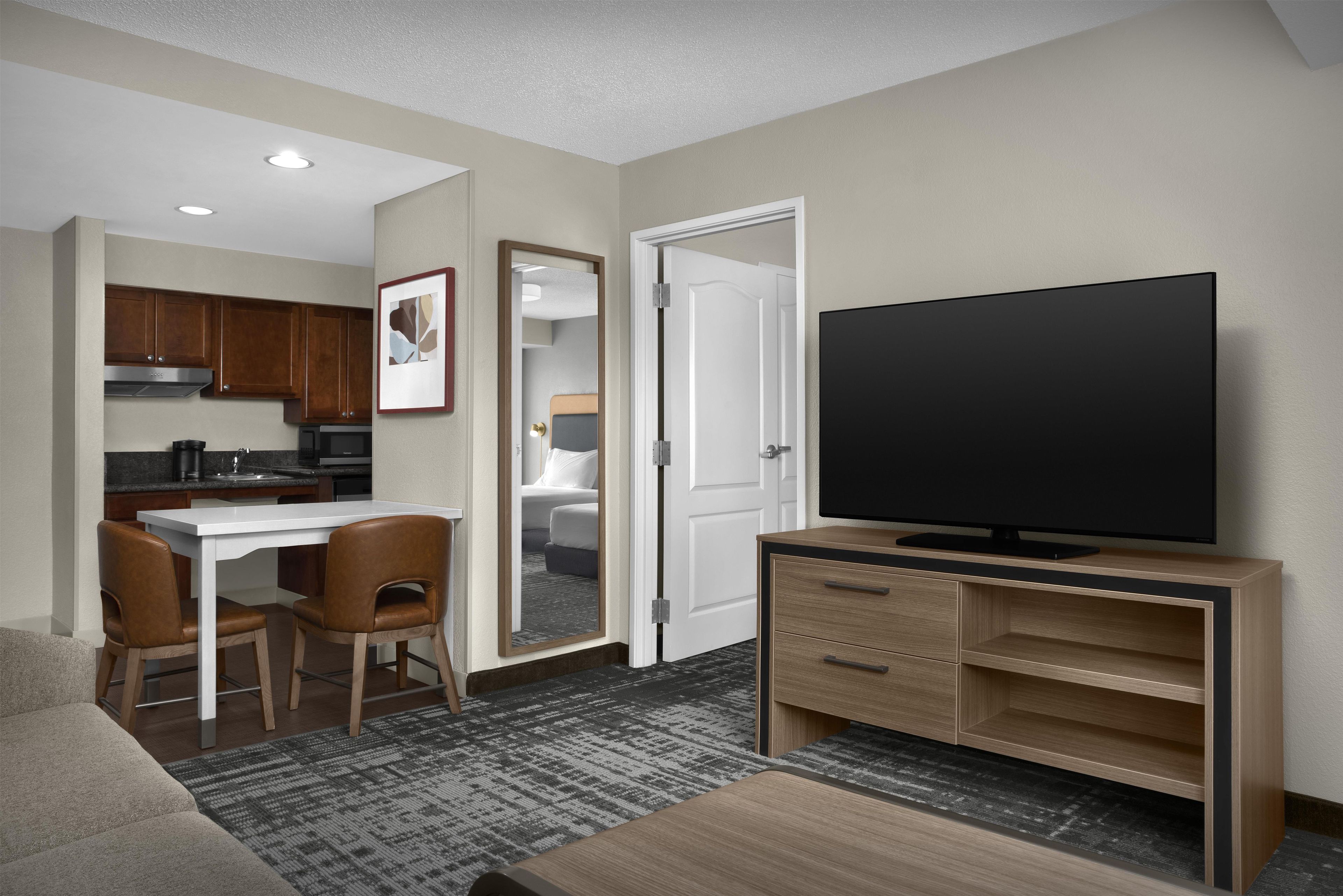 A hotel room with a kitchenette, a dining area with a table and chairs, a TV on a wooden stand, and a door leading to a bathroom.