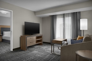 A hotel room with a flat-screen TV on a wooden stand, a desk with a chair, a sofa, and a large window with curtains. - Homewood Suites by Hilton Tampa-Brandon (Tampa)