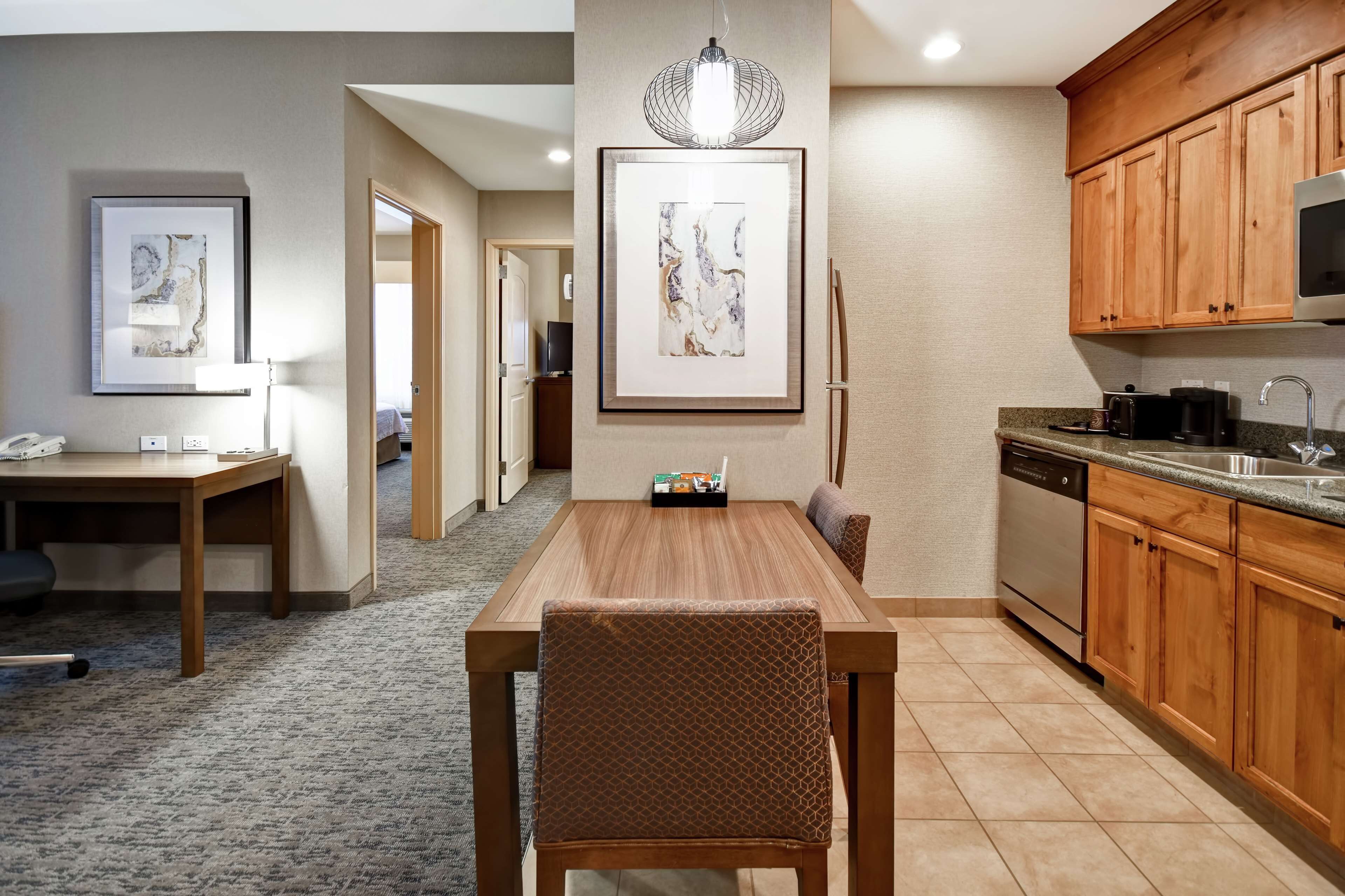 Una habitación de hotel moderna con una mesa de comedor de madera, sillas, una cocina con armarios de madera, horno empotrado y una obra de arte grande enmarcada en la pared.