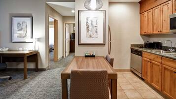 Une chambre d’hôtel moderne avec une table à manger en bois, des chaises, un coin cuisine doté de meubles en bois, un four encastré et une grande œuvre d’art encadrée au mur.