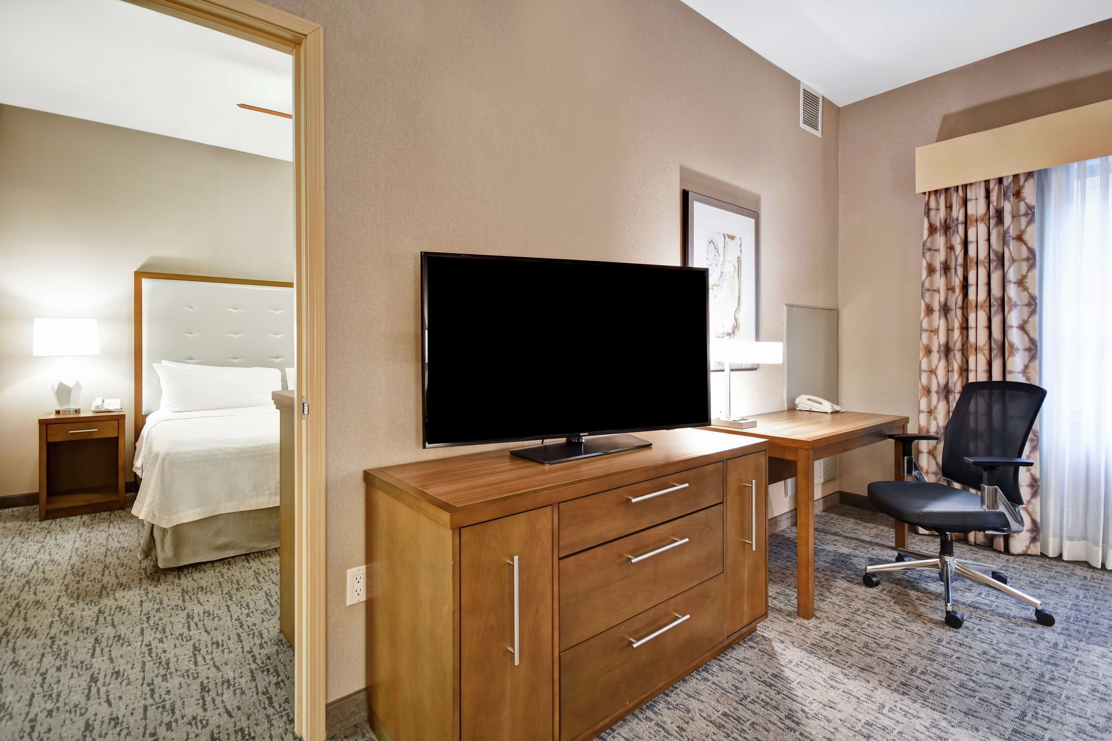 A hotel room with a bed, a TV on a wooden cabinet, a desk with a chair, and a window with curtains.