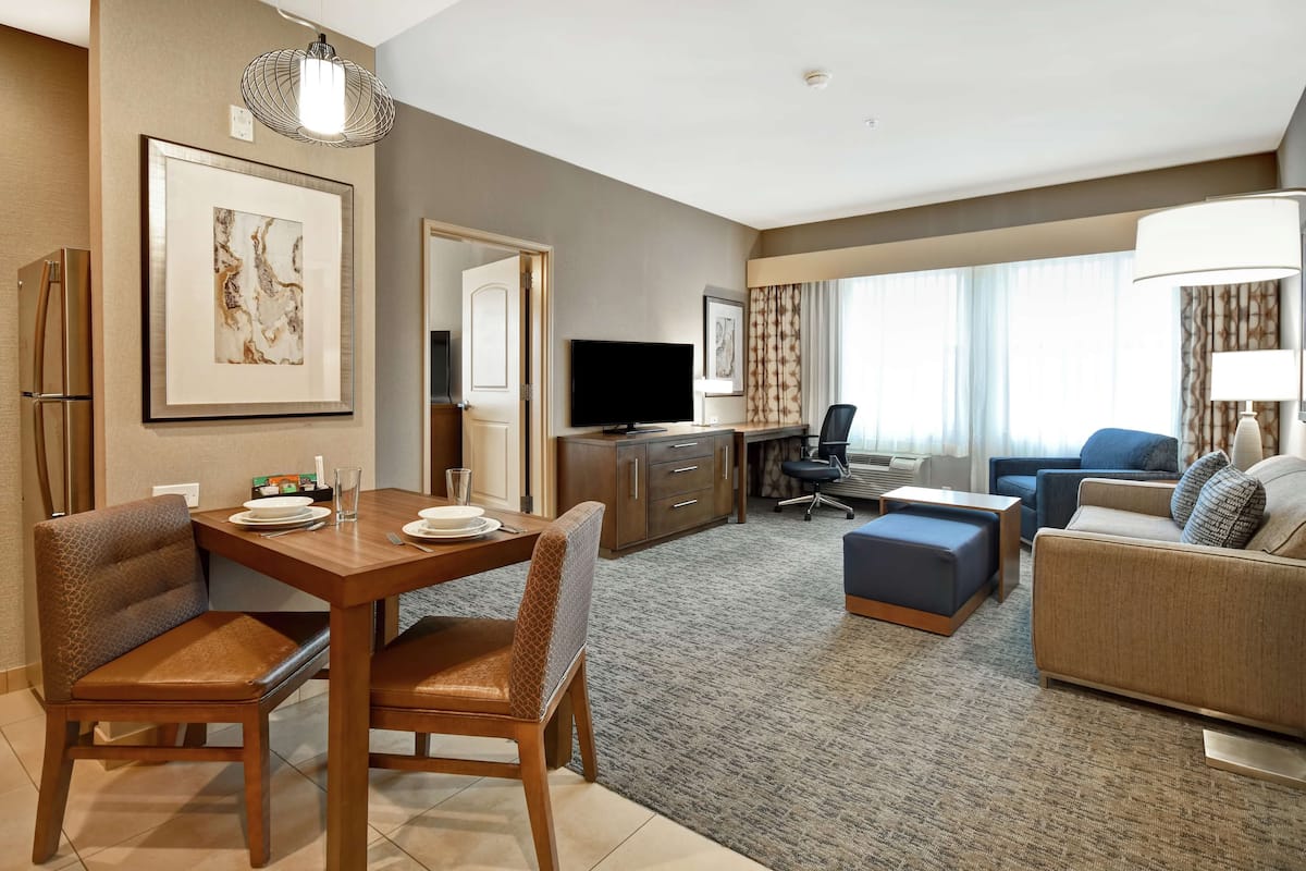 Een hotelkamer met een zithoek, een woonkamer met een bank en fauteuil, een televisie en een groot raam met gordijnen.