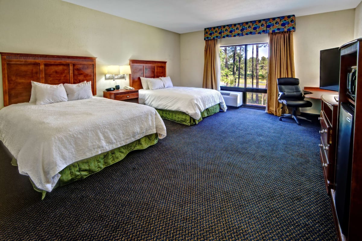 A hotel room with two beds, a desk, a chair, a television, and a large window with curtains.