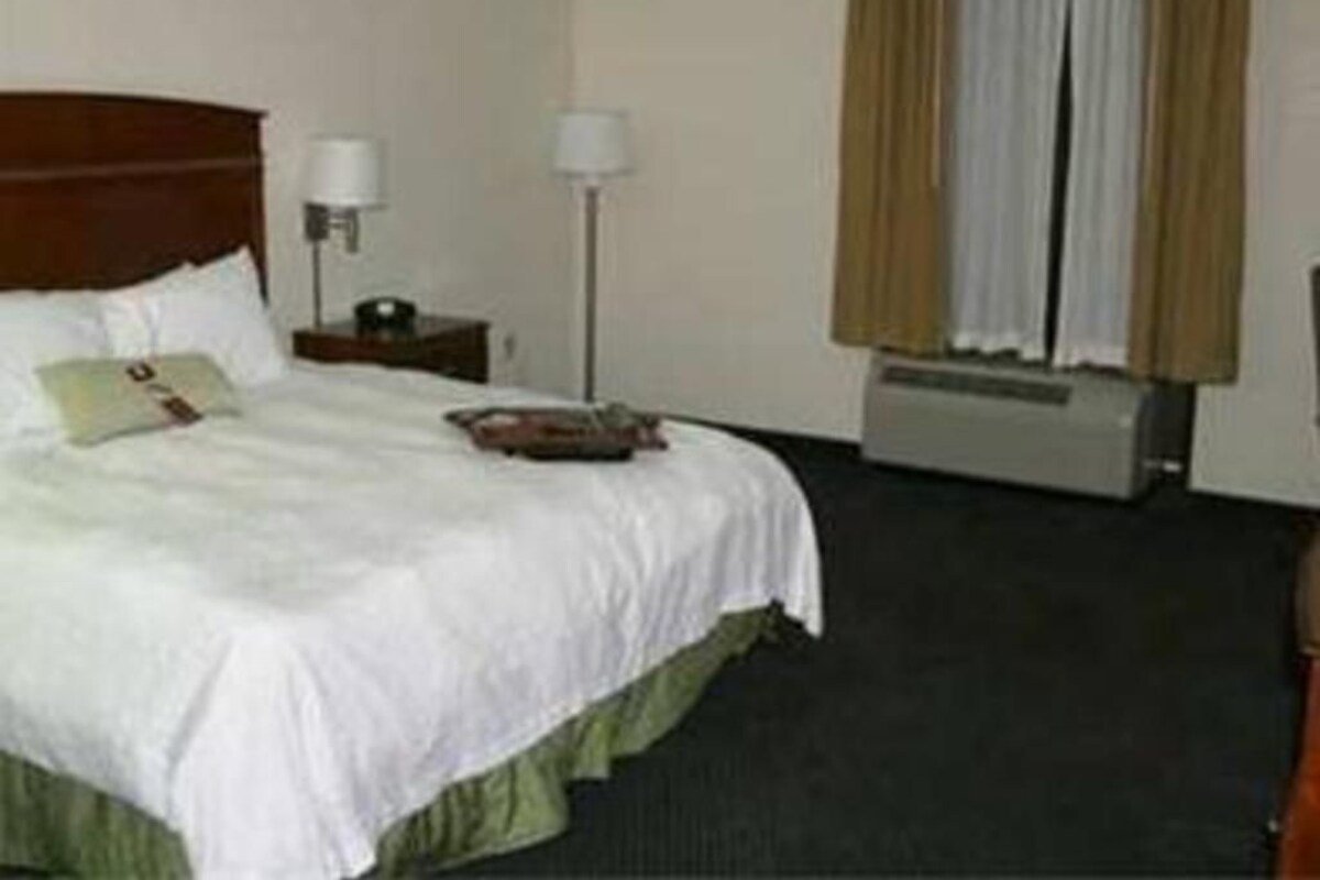Una habitación de hotel con cama, mesita de noche, lámpara y ventana con cortinas.