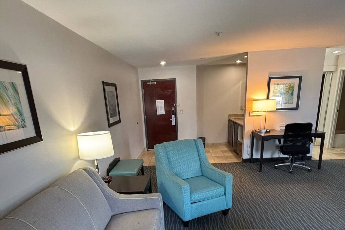 Una habitación de hotel con un sofá gris, un sillón azul, una mesita y un escritorio con una lámpara.
