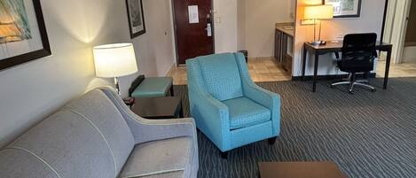 A hotel room with a grey sofa, a blue armchair, a small table, and a desk with a lamp.