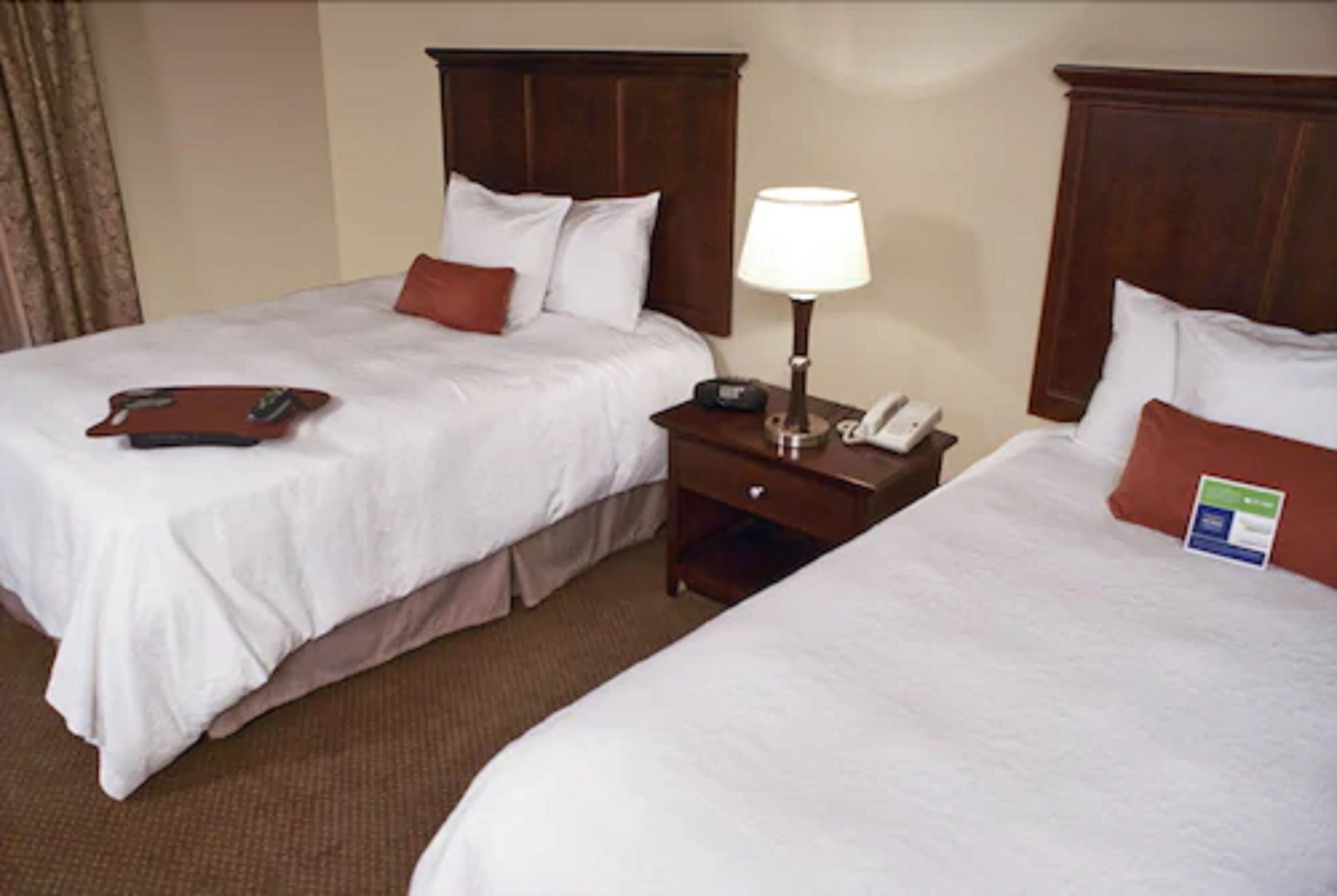 A hotel room with two single beds, each with a wooden headboard, white bedding, and a bedside table with a lamp and a phone.