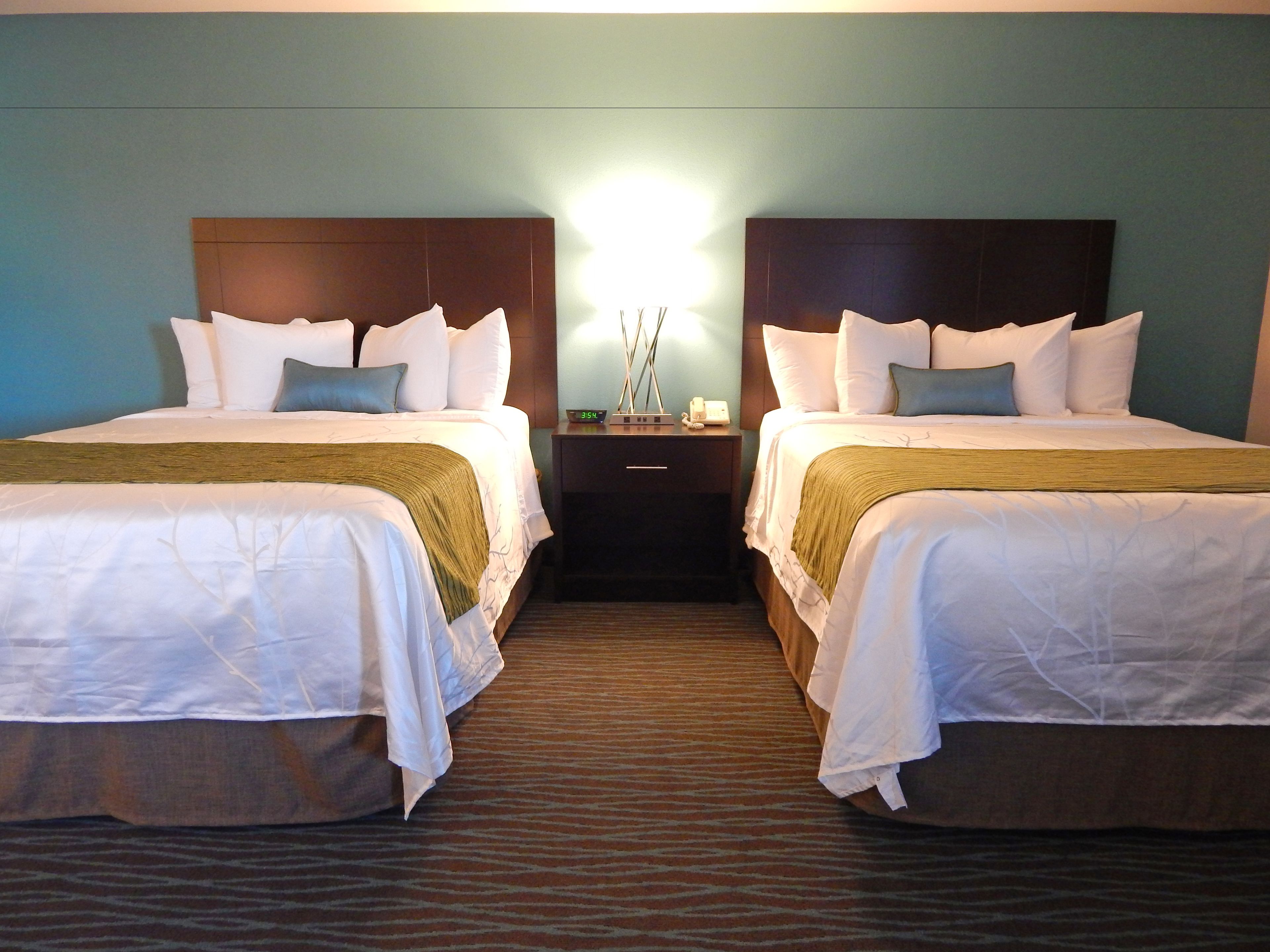 A hotel room with two beds, each with white bedding and a brown headboard. There is a nightstand between the beds with a lamp and a clock.