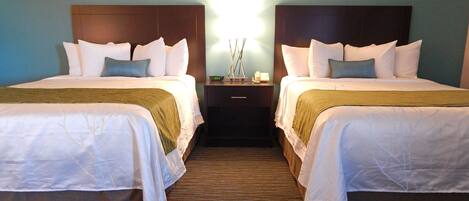 A hotel room with two beds, each with white bedding and a brown headboard. There is a nightstand between the beds with a lamp and a clock.