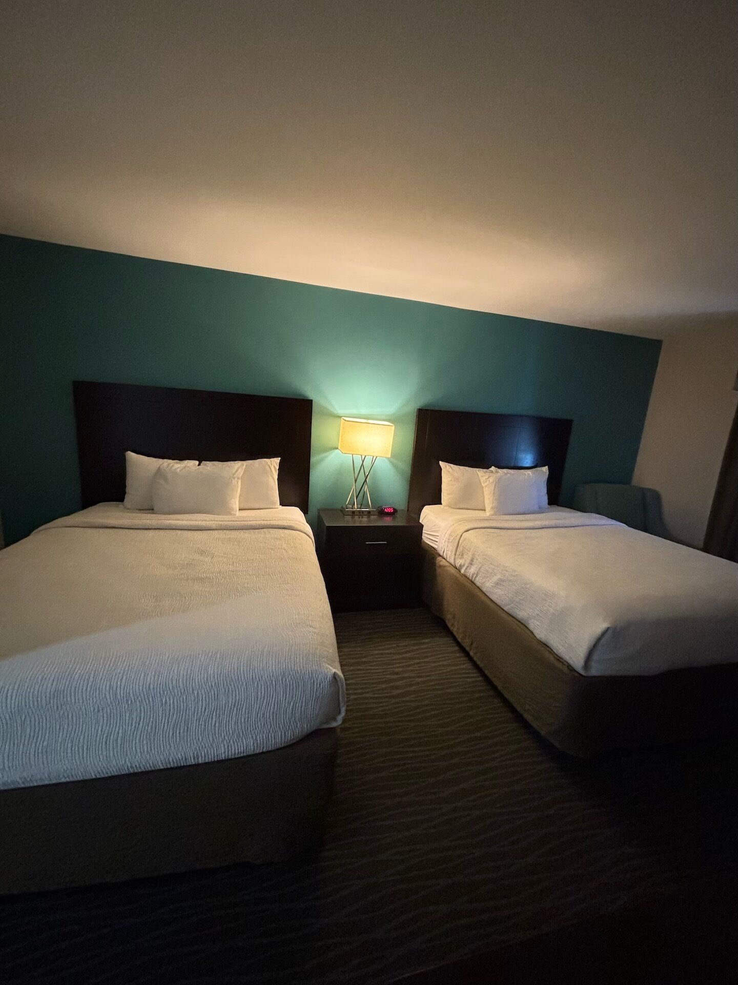 A hotel room with two beds, a nightstand with a lamp, and teal walls.