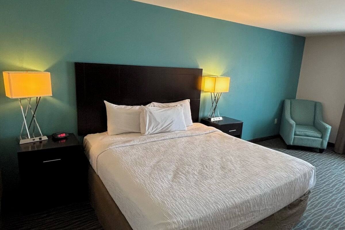 Habitación de hotel con una cama grande, dos mesitas de noche con lámparas, una silla y paredes color verde azulado.