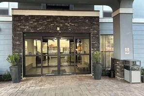 Une entrée d’immeuble avec une façade en pierre, de grandes portes vitrées et deux plantes en pot.