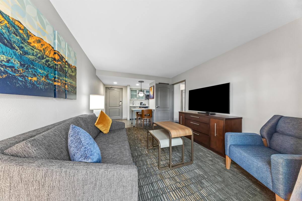 Une chambre d’hôtel avec un canapé gris, un fauteuil bleu, une télévision, une petite table et un tableau au mur.