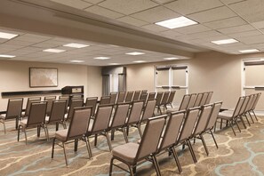 Une salle de conférence avec des rangées de chaises, un sol recouvert de moquette, un écran de projection et une fenêtre avec des rideaux.