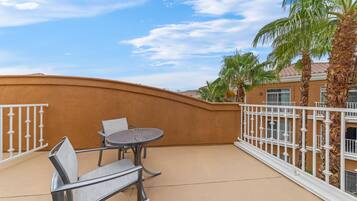 A balcony with a round metal table and two chairs, a white metal railing, and a palm tree.