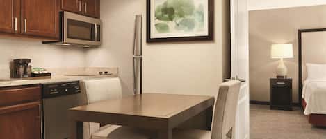 A hotel room with a kitchen area, a dining table with chairs, a bed, a bedside table with a lamp, and a framed artwork on the wall.