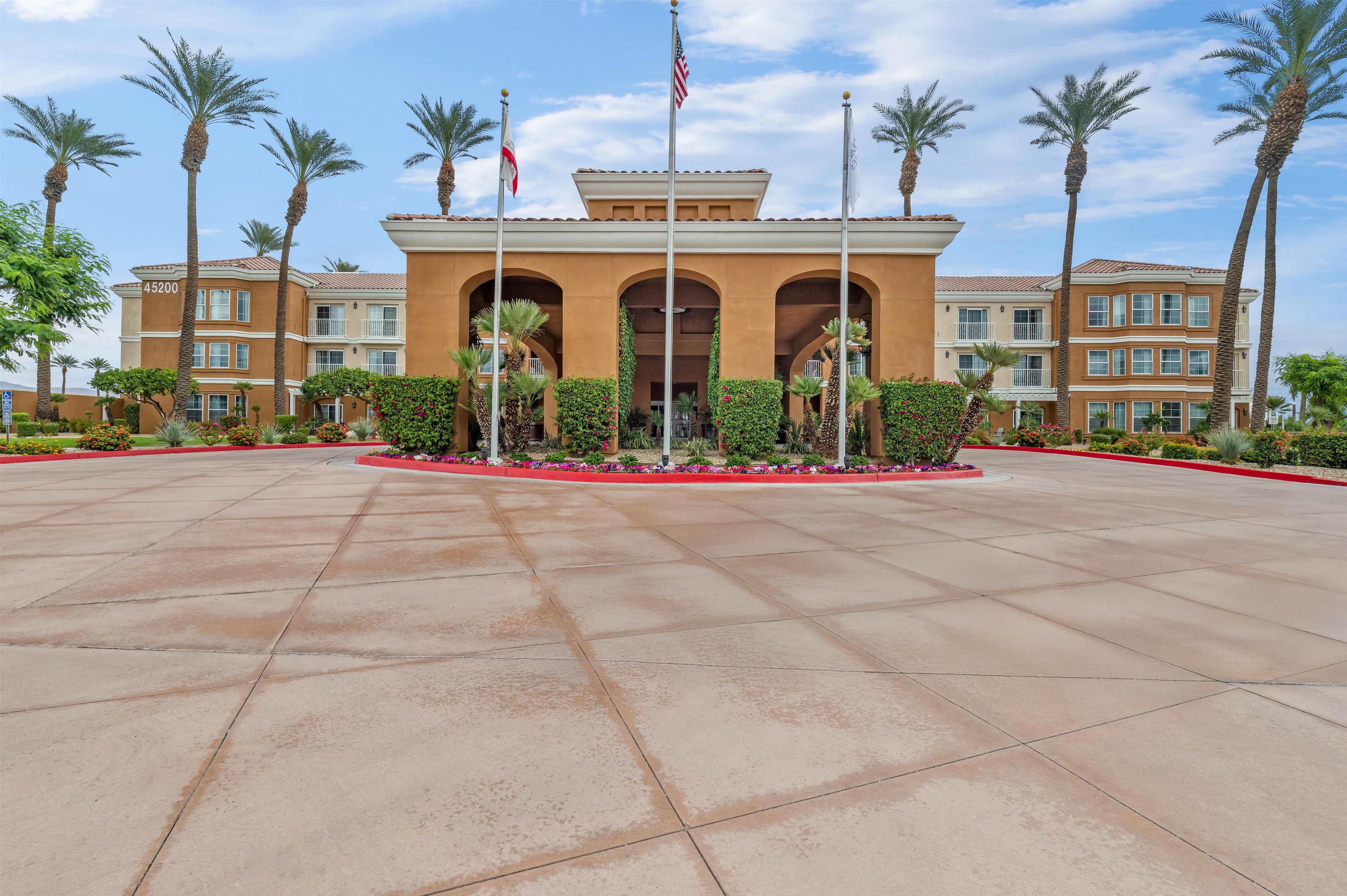 Ingresso di un hotel con un'ampia area pavimentata, palme e un edificio con numerosi balconi.