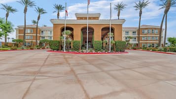 Ingresso di un hotel con un'ampia area pavimentata, palme e un edificio con numerosi balconi.