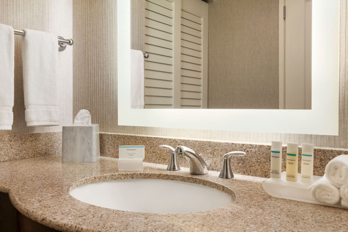 Une salle de bain avec un comptoir en granit, un lavabo blanc, deux robinets, un miroir et des articles de toilette.