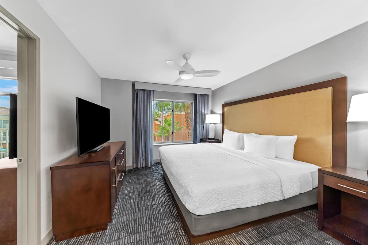 Une chambre d’hôtel équipée d’un lit, d’une télévision, d’un ventilateur de plafond, d’une fenêtre avec des rideaux et offrant une vue sur l’extérieur.